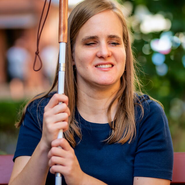 Eine junge Frau sitzt auf einer Parkbank und hält einen Langstock in der Hand; sie hat die Augen geschlossen und lächelt leicht. Im Hintergrund sind Bäume zu sehen.