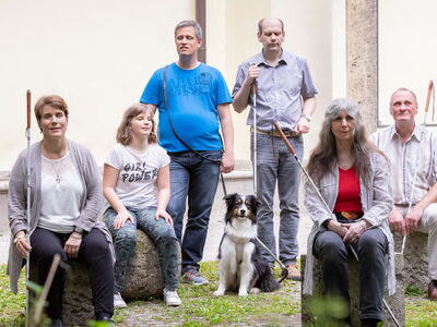 Sechs sehbehinderte und blinde Menschen sitzen und stehen gemeinsam in einem grünen Innenhof. Vier von ihnen halten einen Blindenlangstock, eine Person hat einen Blindenführhund bei sich.