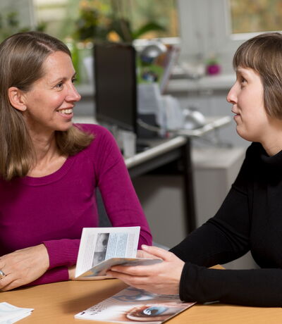Zwei Frauen sitzen an einem Tisch in einem Büro und führen ein Beratungsgespräch; eine von ihnen hält eine Broschüre in der Hand.