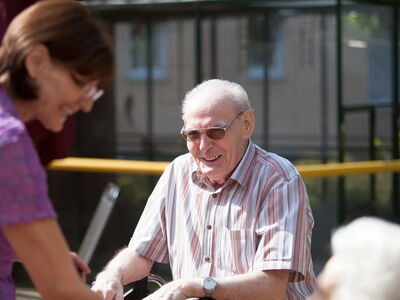 Ein älterer, sehbehinderter Mann mit getönter Brille sitzt draußen vor einem gebäude. Er befindet sich im Gespräch mit einer Frau, die ihm gegenübersteht. Beide Personen lächeln.